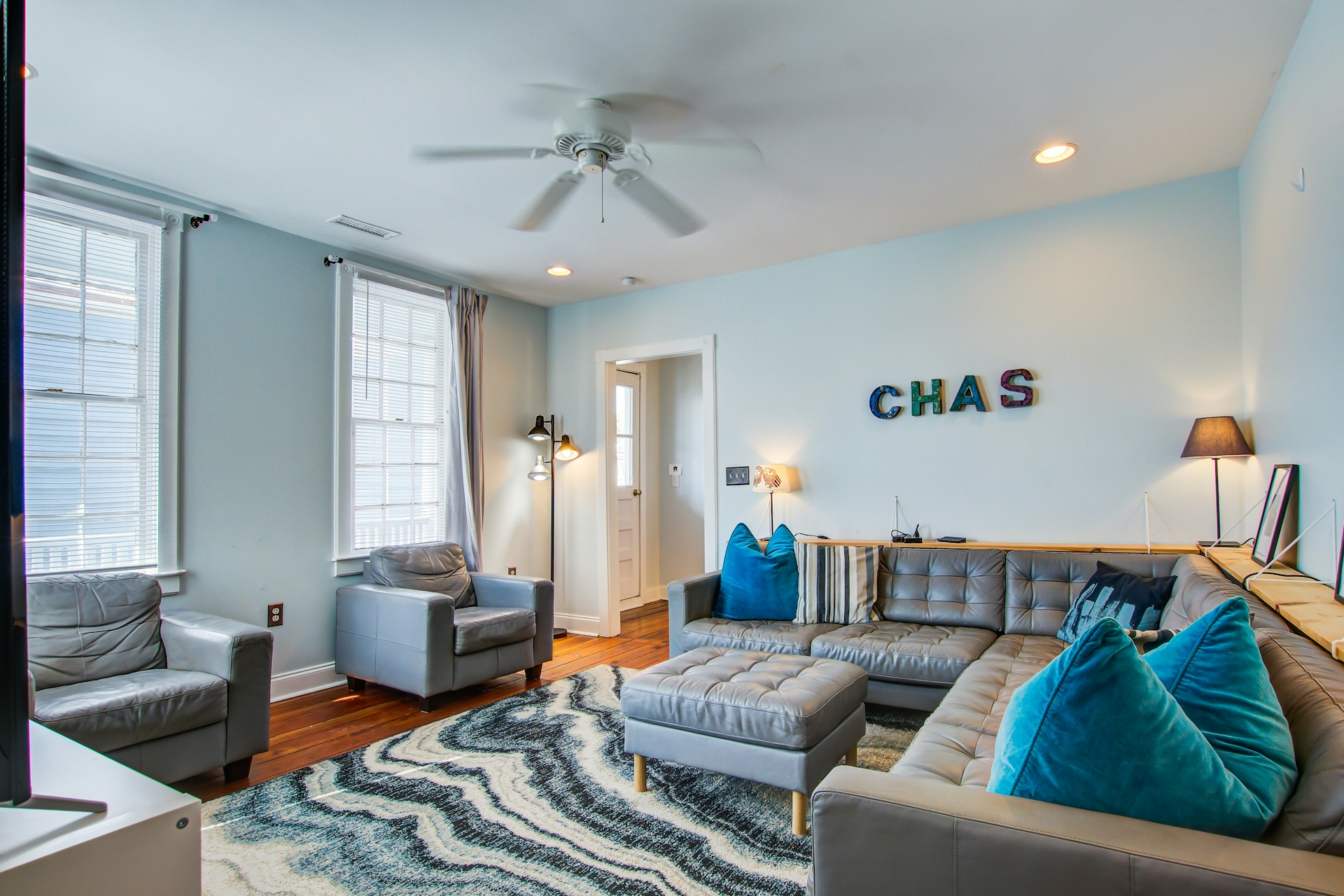 Main living room with gray leather sectional, blue and teal velvet pillows, ottoman, gray wave-pattern rug, ceiling fan, two tall windows with sheer curtains, and CHAS mosaic letter wall art.