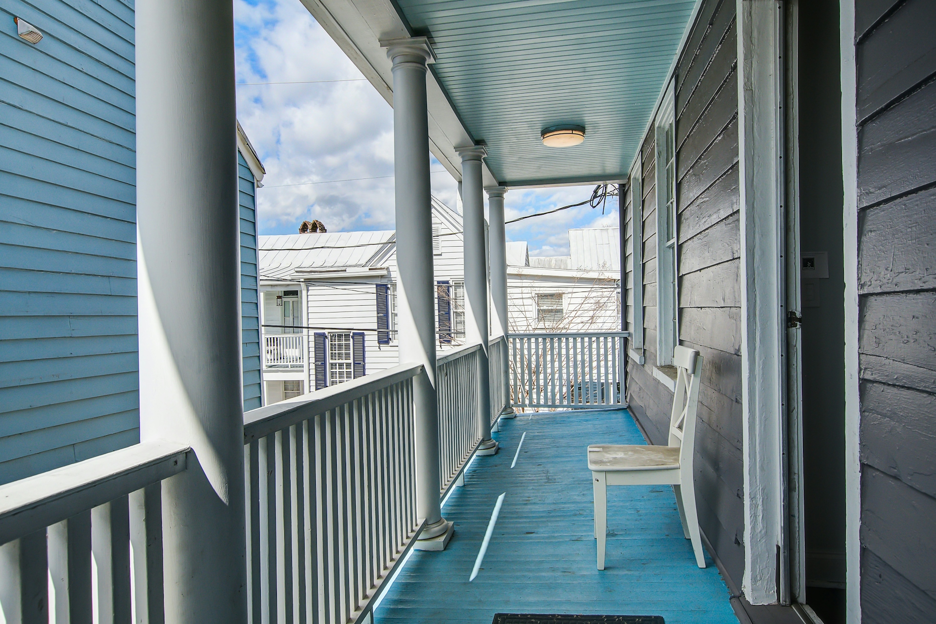 Upper covered porch with sky-blue painted floor and ceiling, white columns and railing, white wooden chair, and view to neighboring rooflines.