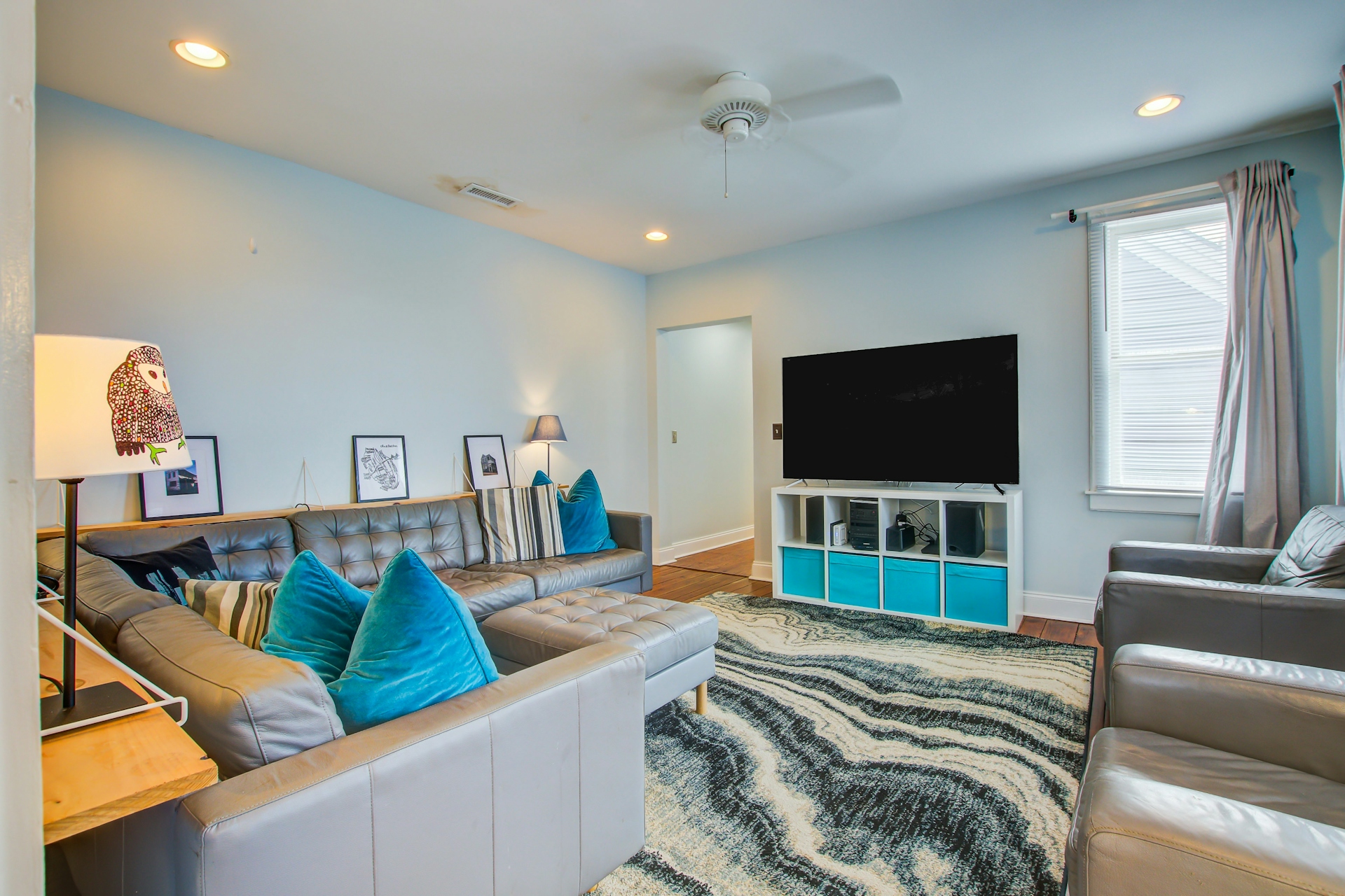 Living room: gray sectional with blue pillows, large TV on white cube media console with teal bin inserts, owl-pattern wall art, recessed lighting, and ceiling fan.