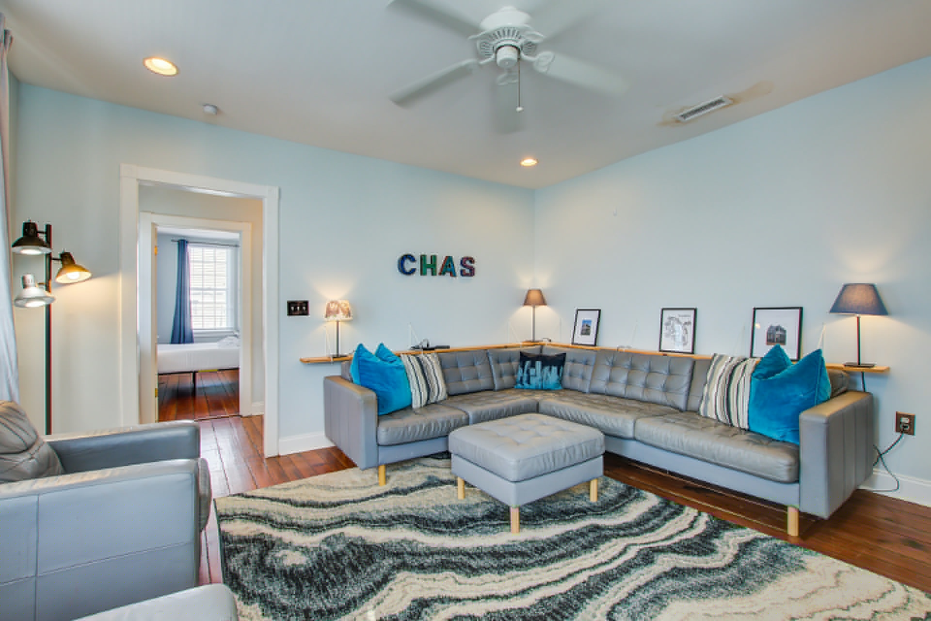 Wide angle of living room showing gray sectional, ottoman, CHAS letters, and open doorway to adjacent bedroom with blue curtains.