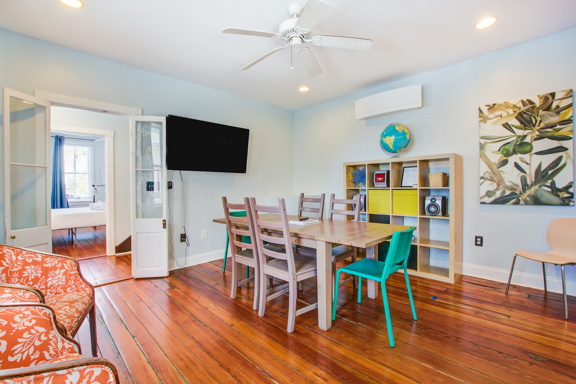 Dining room from opposite end: wall-mounted TV, dining table with chairs, mini-split AC, olive-branch wall art, and French doors into a bedroom.