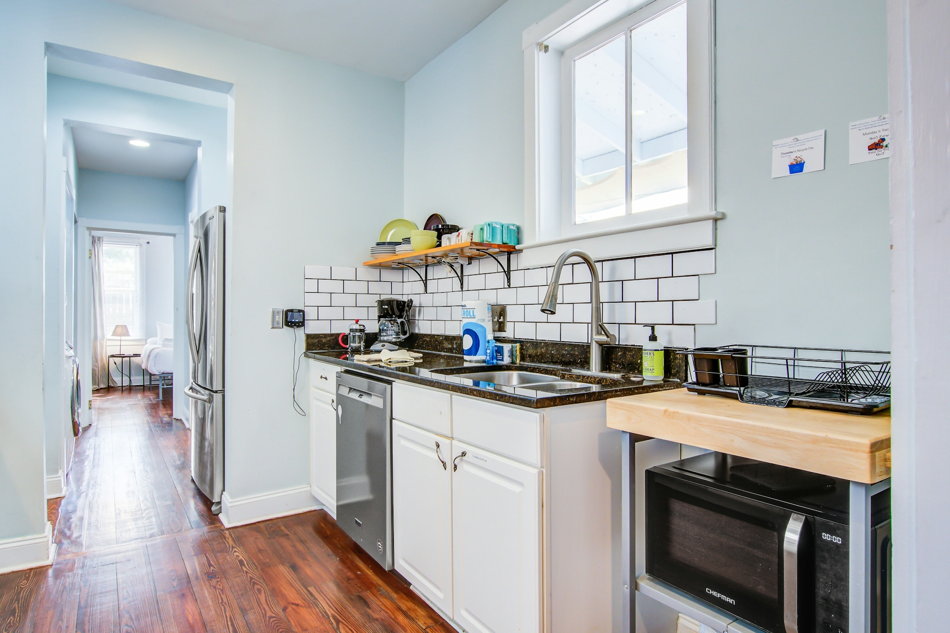 Kitchen with stainless French-door refrigerator, white cabinets, dark granite counters, white subway tile backsplash, double sink under window, dishwasher, and microwave.