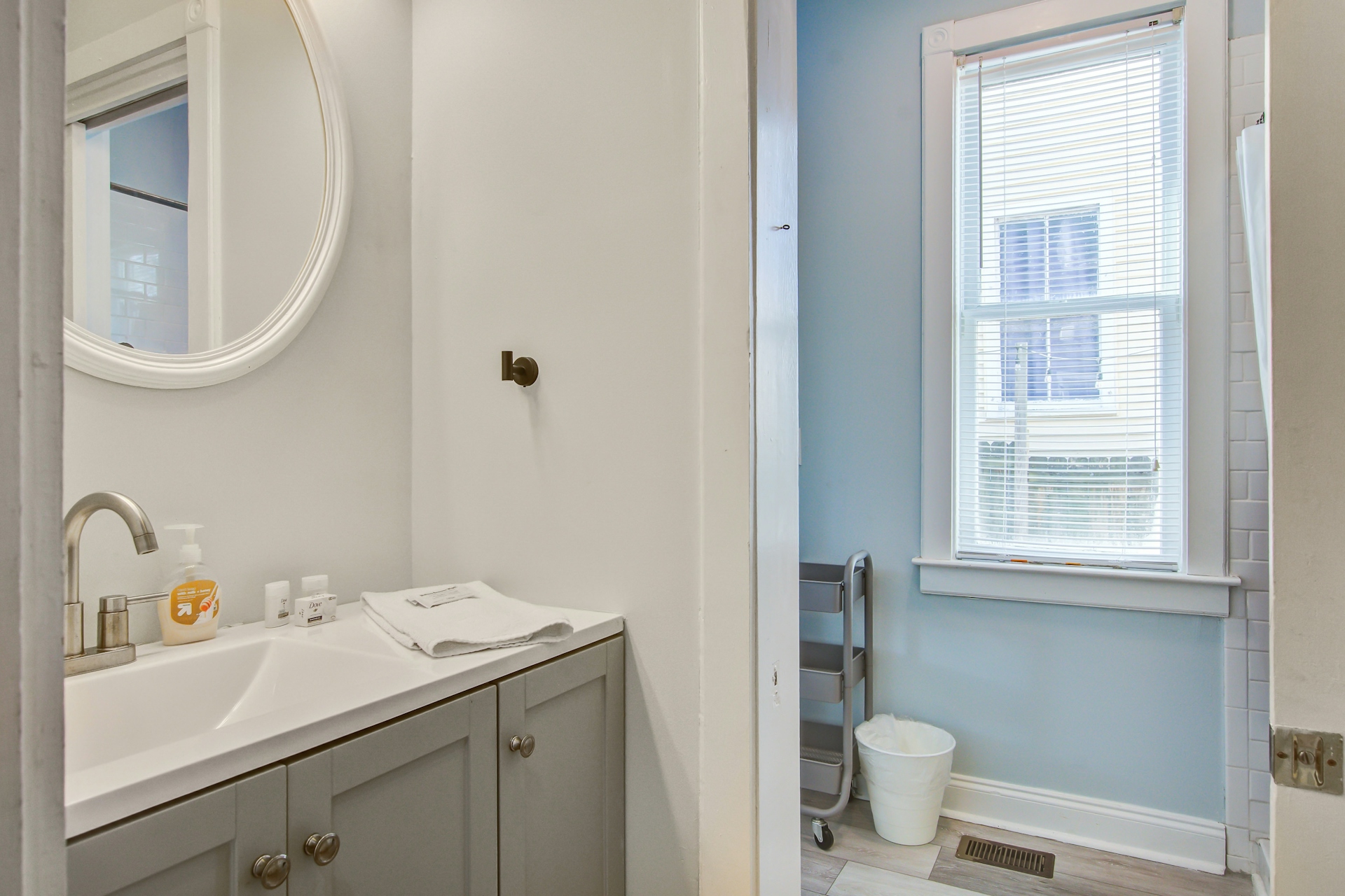 Powder room with gray vanity, white sink, round white-framed mirror, toiletries, and adjacent blue-walled nook with rolling cart and tall window.