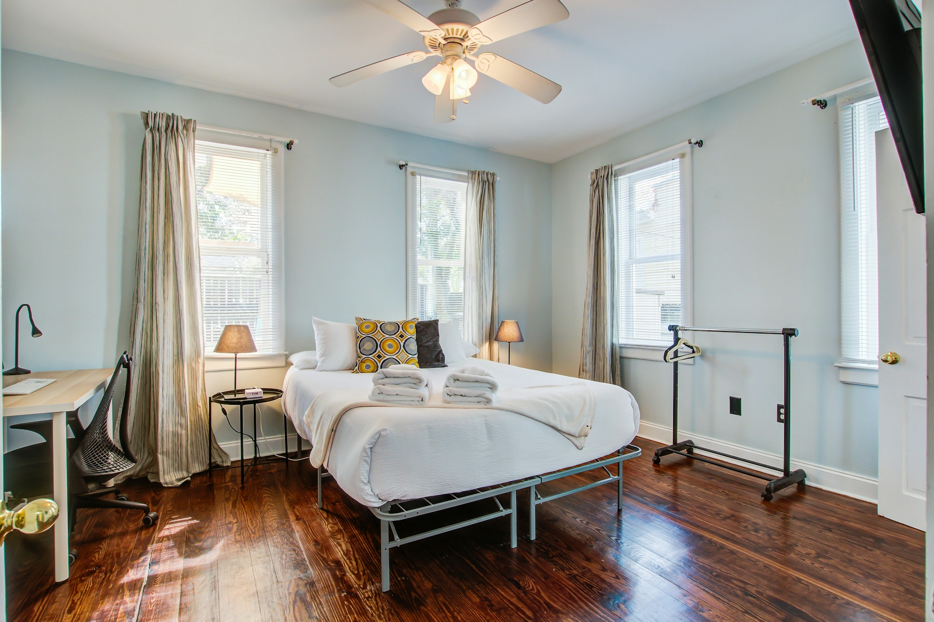Bright bedroom with queen bed in white linens, three windows with striped curtains, desk and chair, garment rack, and ceiling fan.