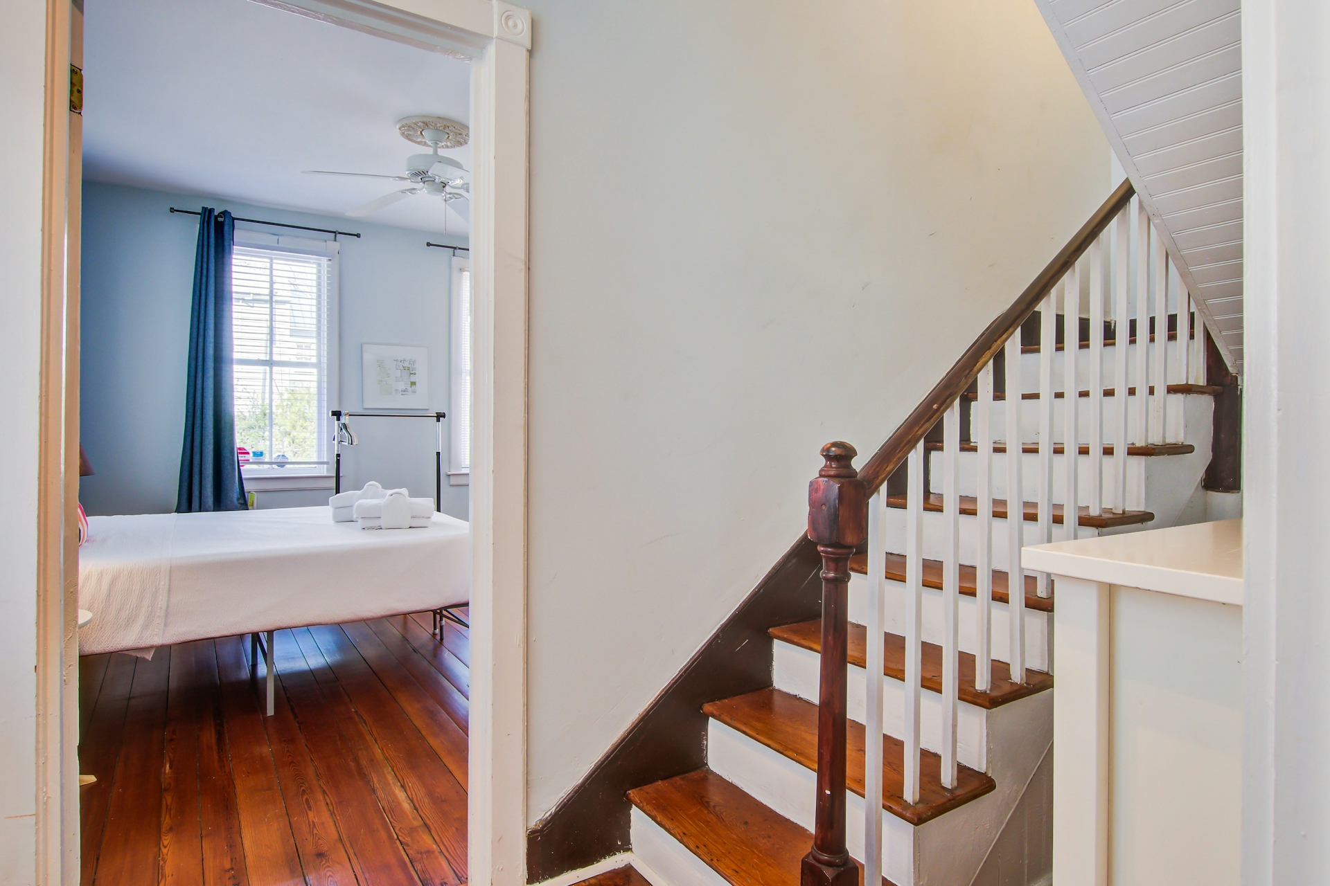 Interior staircase with stained wood treads, white risers and balusters, dark handrail, and doorway to bedroom with blue curtains.
