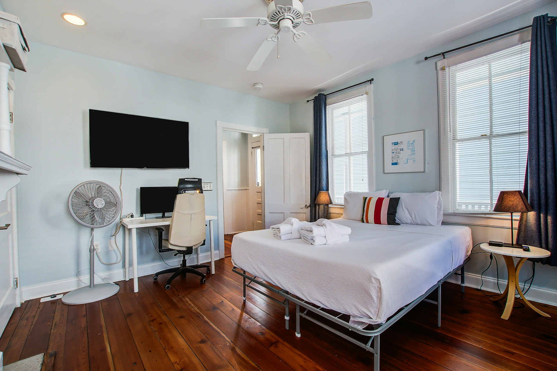 Bedroom with wall-mounted TV, queen bed in white, desk with monitor and office chair, standing fan, blue curtains, and pine plank floors.