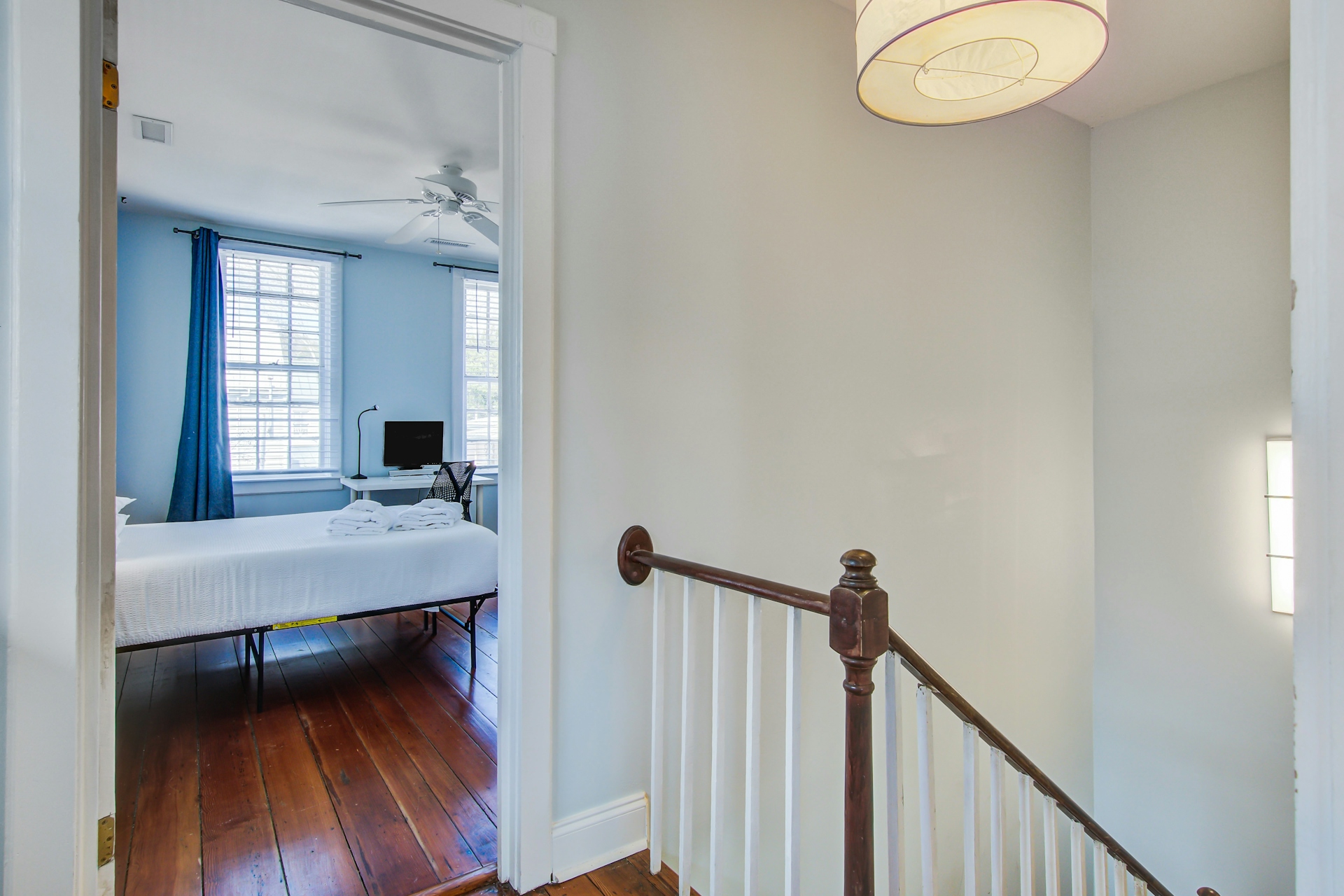 Upstairs landing with drum-shade pendant, white walls, dark stair railing, and view through doorway to bedroom with blue curtains.