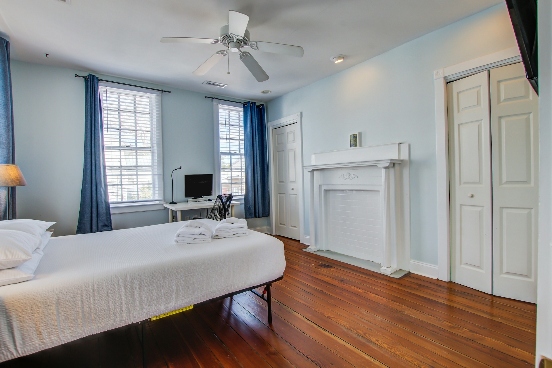 Bedroom with decorative white fireplace mantel, queen bed, desk with monitor, blue curtains over two windows, and louvered closet doors.