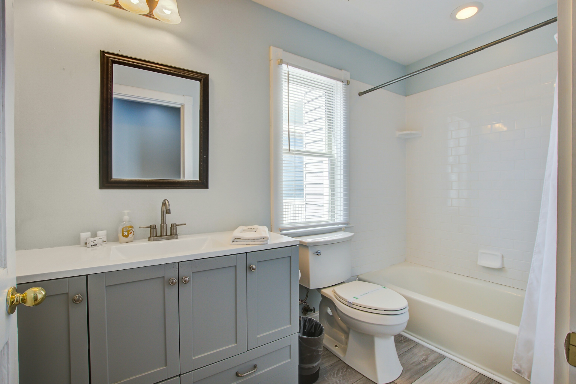 Full bathroom with gray vanity, white quartz countertop, framed mirror, tall window, and white tub-shower combo with subway tile surround.