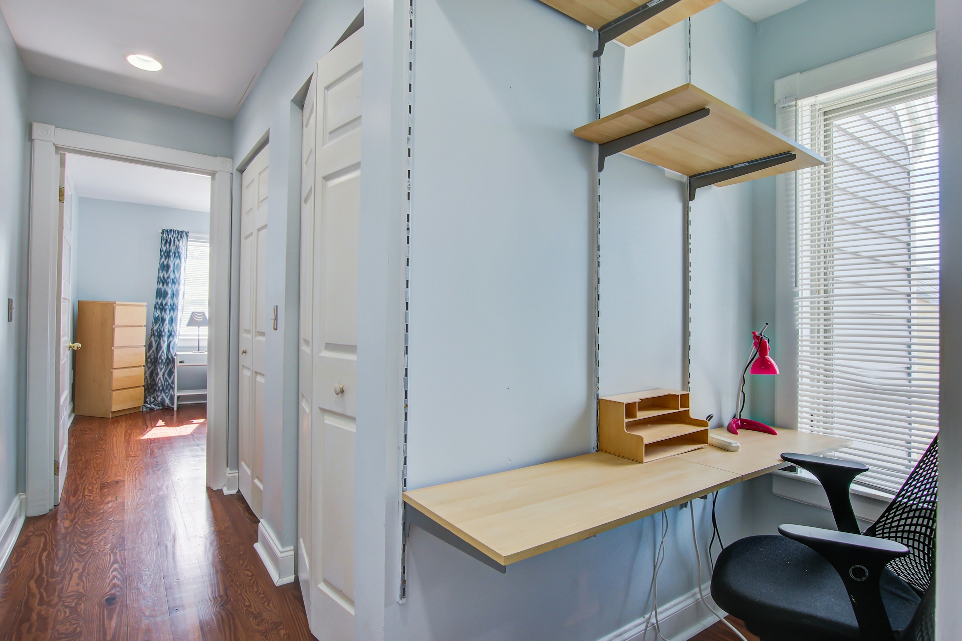Hallway nook with built-in desk and shelving, ergonomic chair, pink desk lamp, closet doors, and view toward bedroom at the end.