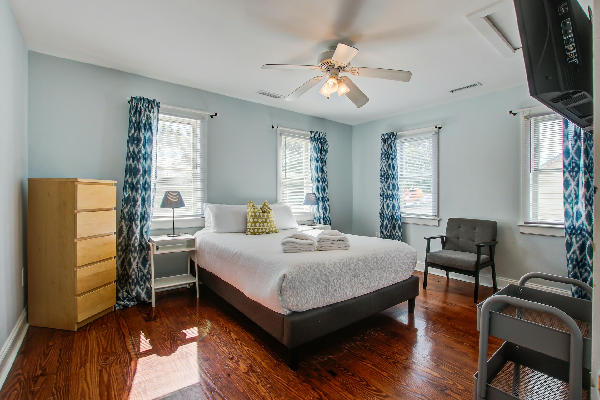 Bedroom with queen bed, wooden dresser, gray accent chair, three windows with blue ikat-pattern curtains, ceiling fan with light, and pine floors.