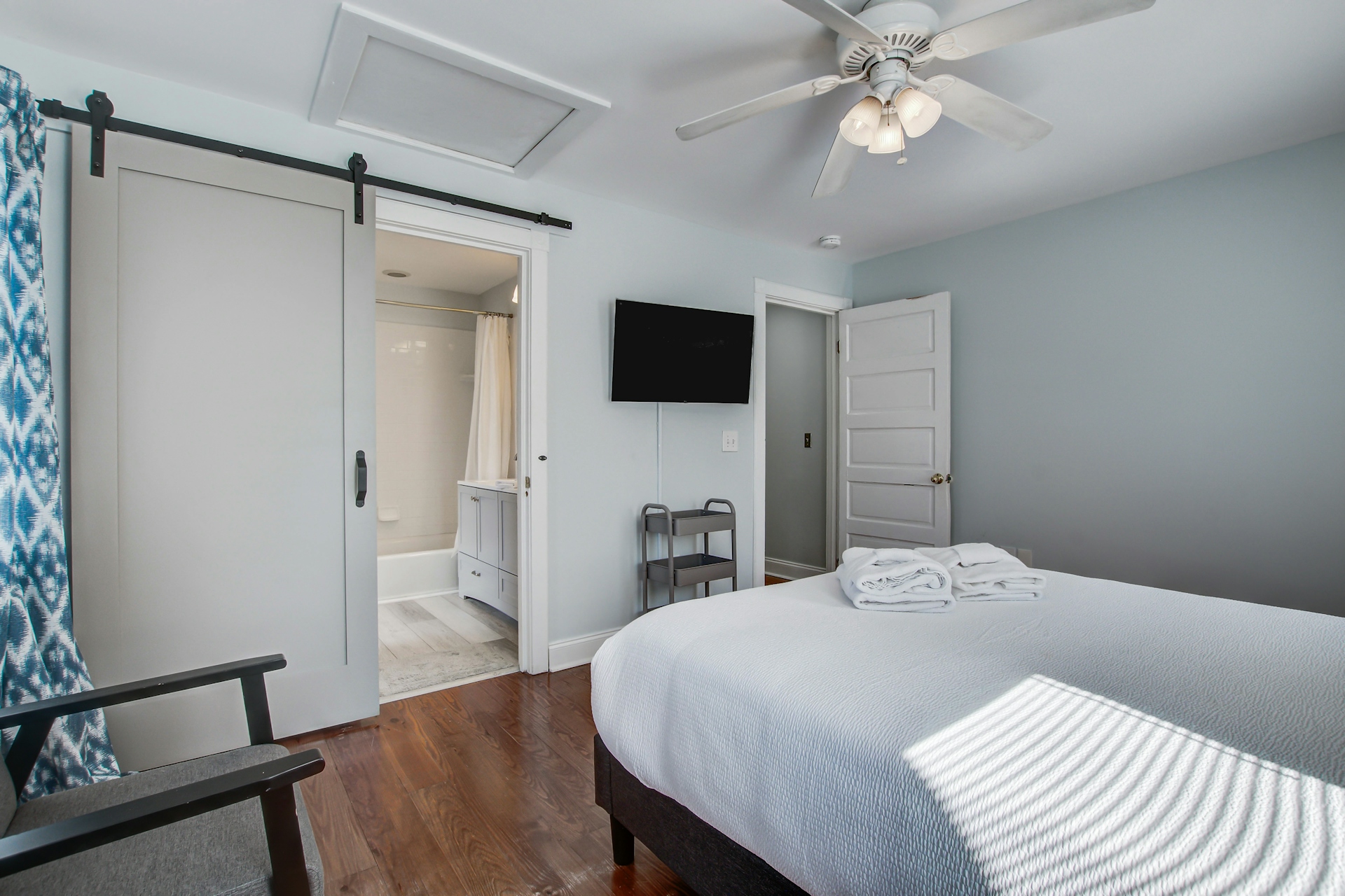 Bedroom with sliding barn door to ensuite bathroom, queen bed, wall-mounted TV, gray armchair, attic access, and ikat-pattern curtains.