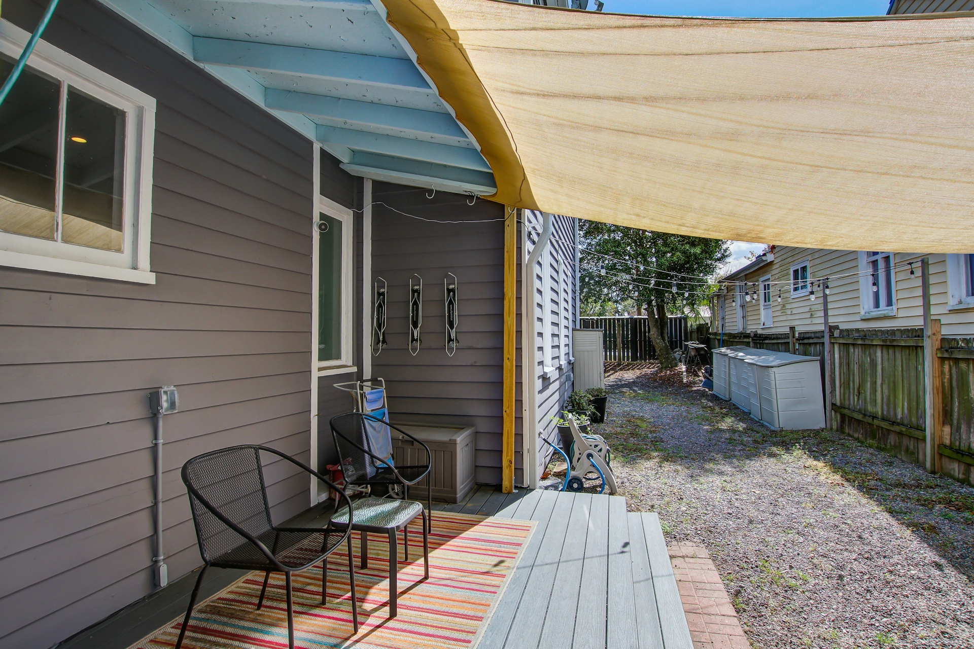 Side porch with beige shade sail, black metal chairs, side door, and gravel yard with neighboring shed visible.